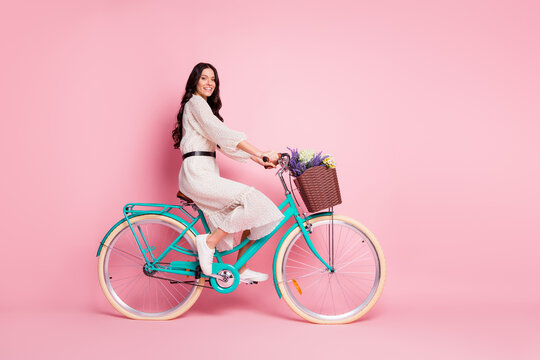 Full length body size photo of pretty woman riding bike with wild flowers in white dress isolated on pastel pink color background