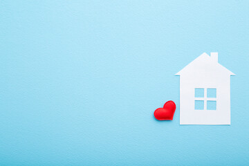 White paper house with bright red heart on light blue table background. Pastel color. Closeup. Empty place for text. Top down view.