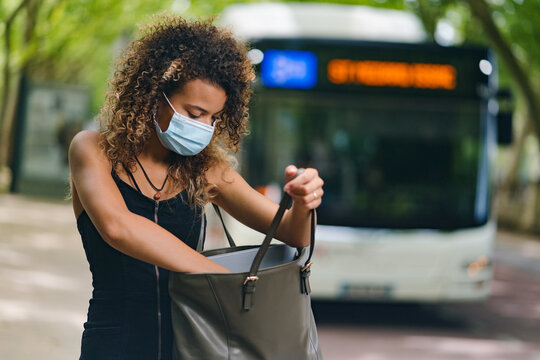young woman taking smart phone from handbag at the bus station and wearing a surgical mask