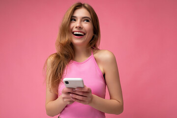 Photo of smiling happy good looking young blonde woman wearing pink top poising isolated on pink background with empty space holding in hand and using mobile phone messaging sms looking up and