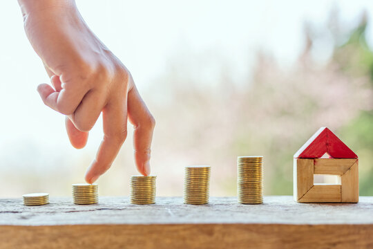 House model resting on wooden desk and hand walking on coin stack