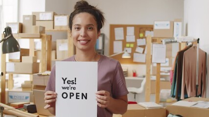 Portrait of young joyous mixed-race woman holding poster with Yes! WeвЂ™re open sign and smiling at camera while standing in office