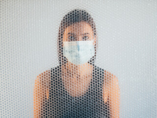 Air pollution. Defocused portrait. Respiratory disorder. Concerned woman in protective face mask behind transparent polyethylene bubble wrap texture drop on light background out of focus.