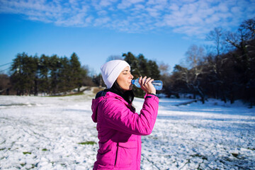 Female athlete in winter clothes drinking water before training on snow. Jogging and recreation.