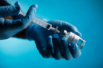 mani di un medico con guanti in lattice, prelevano il vaccino anti-covid con una siringa da una filetta di vetro, isolato su sfondo blu - obrazy, fototapety, plakaty