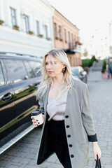 Obraz premium modern young blonde in the city is walking against the background of a car with a cup of coffee.