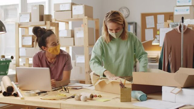 Young woman in protective mask working on laptop in office of online clothes store while her female colleague packing shirt into box before shipping order to customer during coronavirus outbreak