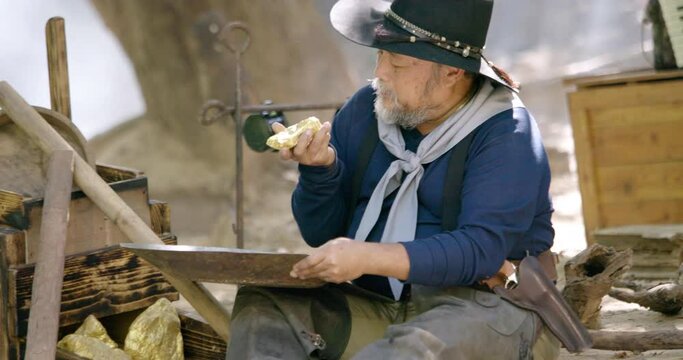 Gold digger wearing on cowboy hat and cowboy boots sitting using gold panning to refining the gold metal particles  while boiling the coffee flask above fire and some are play revolver gun