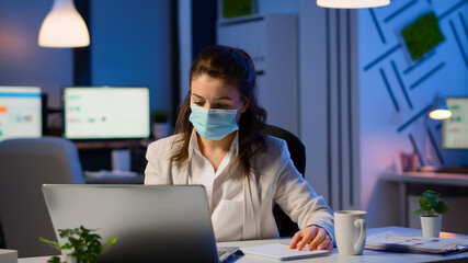 Employee with protection face mask working late at night in new normal business office to respect deadline of project, taking notes, analysing documents sitting at desk overtime during global pandemic