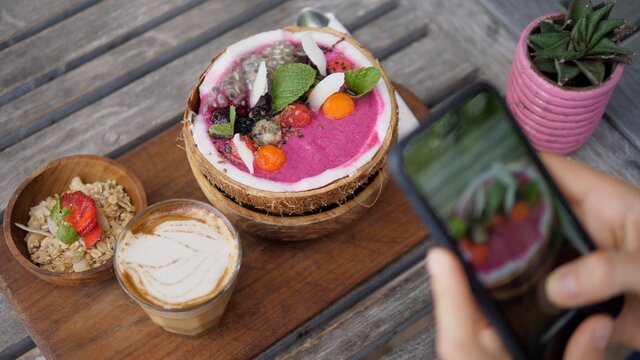 Top view of vegan breakfast of smoothie coconut bowl served on wooden board with dairy free latte being photographed with a smartphone 