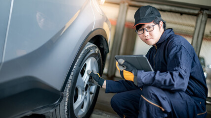 Asian auto mechanic holding digital tablet checking car wheel and tire in auto service garage. Mechanical maintenance engineer working in automotive industry. Automobile servicing and repair concept