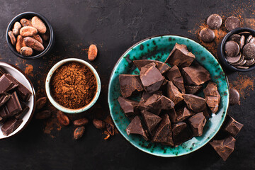 Pieces of broken dark chocolate in a bowl and ingredients for chocolate sweets on a dark textured background, top view. Confectionery chocolate background. Chunks of chocolate.