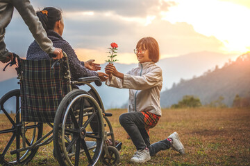 Daughter giving a rose flower her disabled handicapped woman sitting on her wheelchair in sunset...