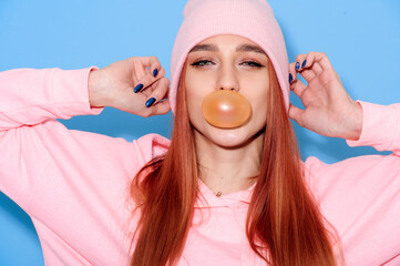 Young attractive girl in pink clothes blowing bubbles from chewing gum on blue background
