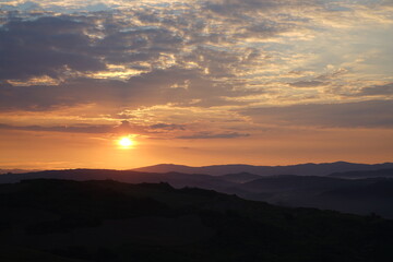 tramonto , toni caldi , con sole tra strati di nubi in alto e susseguirsi di montagne in basso