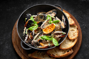 Cooked mussels in a skillet with cheese and basil leaves, on a wooden board, on a dark background