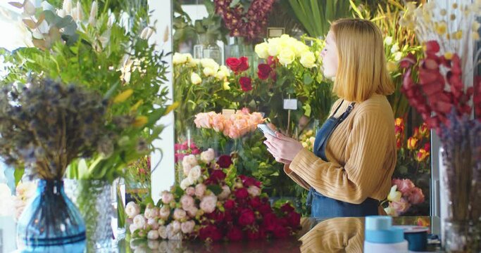 Medium shot of woman working in flower store, using smartphone. Pretty female florist making blossom bouquet, adding photos to social media using phone. Designing, floral workshop, working concept.