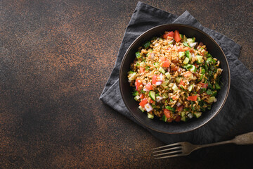 Salad of whole grain cereal spelt with seasonal vegetables, tomato, cucumber in bowl on brown background. Top view. Copy space.
