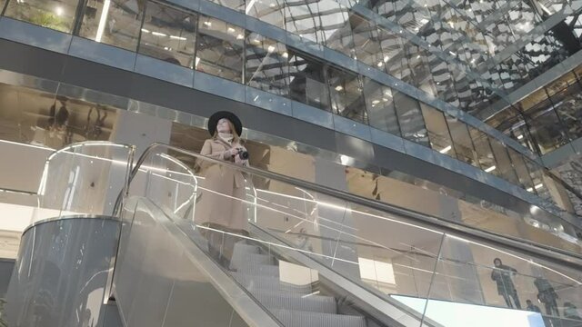 Woman in beige coat and black hat going down the escalator inside the modern shopping center. Action. Young female holding a professional camera and taking pictures in a shopping mall.
