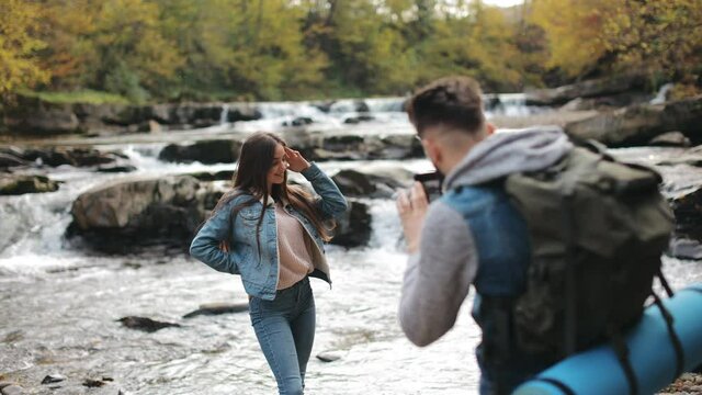 A man is taking pictures of a young woman standing by a beautiful mountain river. Hiking in the mountains. 4K.