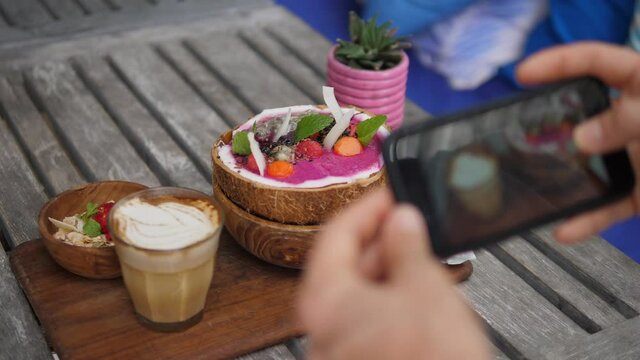 Smartphone picturing tropical smoothie bowl with coffee latte served on wooden tray. Sharing food on social media