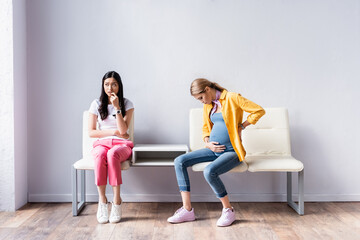 Pregnant woman sitting near worried asian woman in queue