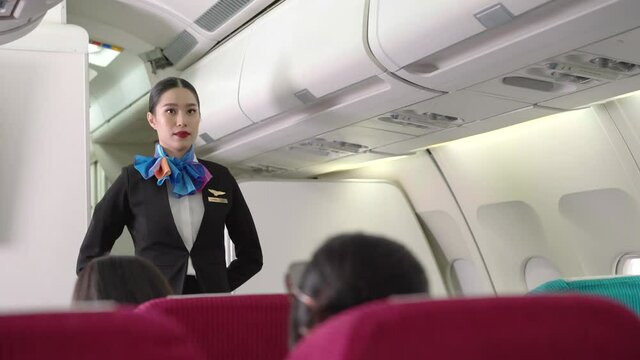 Asian female air hostess standing in  front of cabin seat, watching and checking passenger ready on airplane before takeoff. safety and business security. travel or go to work abroad concept