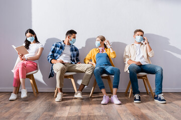 Fototapeta premium Multicultural people in medical masks talking and reading book on chairs in hall