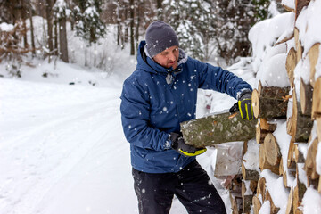 Portrait of smiling handsome young man in winter forest taking logs for his fireplace, copy space. Man arranging wood on snowy yard for a house fireplace with heavy snowflakes background.