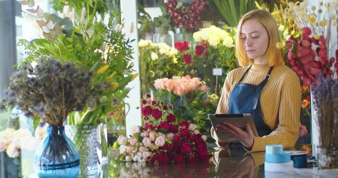Medium shot of woman working in flower store, using tablet. Pretty female florist selling blossom bouquet, making audit using modern gadgets. Designing, floral workshop, working concept.