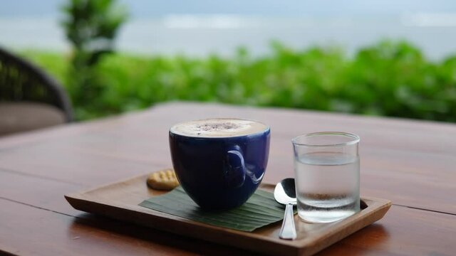 Hands serving espresso macchiato with complimentary cookie on wooden tray. Coffee break with an ocean view