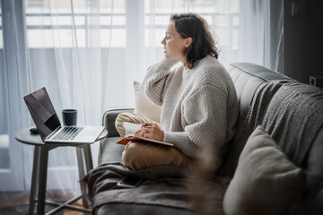 Beautiful young girl student studying at home online while sitting in warm sweater on the couch