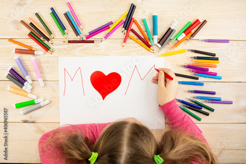Little girl makes a greeting card to mom with a heart for mother's day. Drawing and applique work are done by the child using colored pencils or felt-tip pens. Children's drawing. View from above