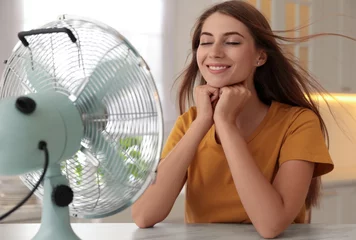 Foto op Plexiglas Woman enjoying air flow from fan at table in kitchen. Summer heat © New Africa