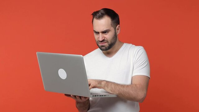 Displeased worried confused bearded young man 20s in white casual t-shirt isolated on orange background studio. People lifestyle concept. Working on laptop pc computer put hand on head spreading arms