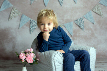 Cute blond toddler child, boy, sitting on little baby armchair, reading a book and holding flowers