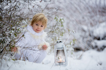 Beautiful blond toddler child, boy with white knitted handmade overall, holding lantern in the snow