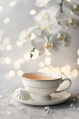 Cozy home spring breakfast still life. A porcelain cup of hot green tea with a marshmallow dessert on a gray background is decorated with a flowering branch of a white otchid. Spring concept. 