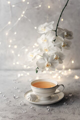 Cozy home spring breakfast still life. A porcelain cup of hot green tea with a marshmallow dessert on a gray background is decorated with a flowering branch of a white otchid. Spring concept. 