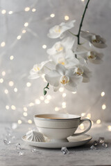 Cozy home spring breakfast still life. A porcelain cup of hot green tea with a marshmallow dessert on a gray background is decorated with a flowering branch of a white otchid. Spring concept. 