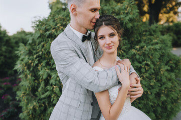 Fototapeta premium Stylish groom in a gray suit and black bow-tie gently hugs a cute bride in a white dress in the park against a background of green plants, thuj. Wedding portrait of smiling newlyweds.
