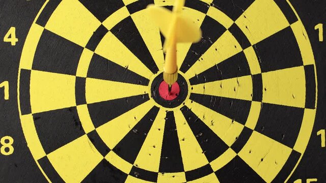 The yellow dart hits the center of the dartboard, then hand removes his, closeup