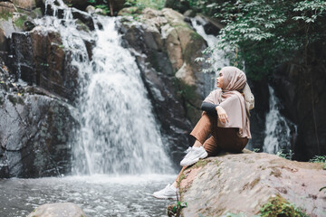 Young asian muslim tourist woman wearing brown hijab sitting on the rock in front of waterfall.