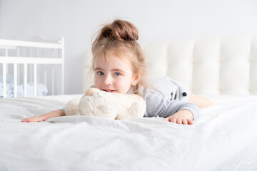 little girl in grey turtleneck playing with teddy bear on white bedding