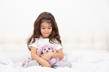Obraz na płótnie Canvas little girl in home wear sits with teddy bunny on bed at home