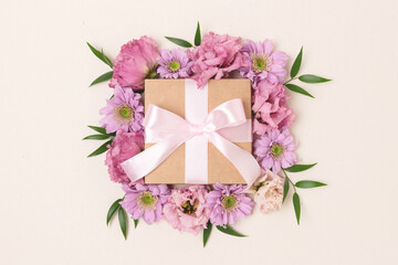 Gift box with a tied ribbon bow and flower frame. Springtime composition on a beige background.