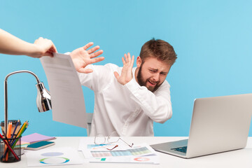 Unhappy bearded man analyst hiding from extra work sitting at workplace with laptop, holding out...