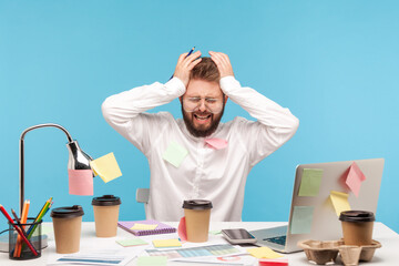 Scared busy man analyst with beard sitting at workplace with coffee cups all covered with sticky notes, has lot of paper work, overworking. Indoor studio shot isolated on blue background © khosrork