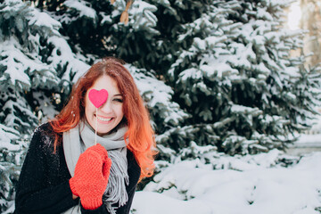 A girl with red hair holds a heart on a stick in her hands against a background of trees and snow. Valentine's day, winter, love, feelings, discounts and promotions.