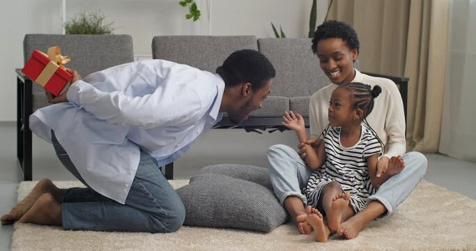 Little cute girl sits with her mother on floor in living room mom covers daughter's eyes with hands, dad black man comes in with present gives baby red gift box for birthday makes surprise for child